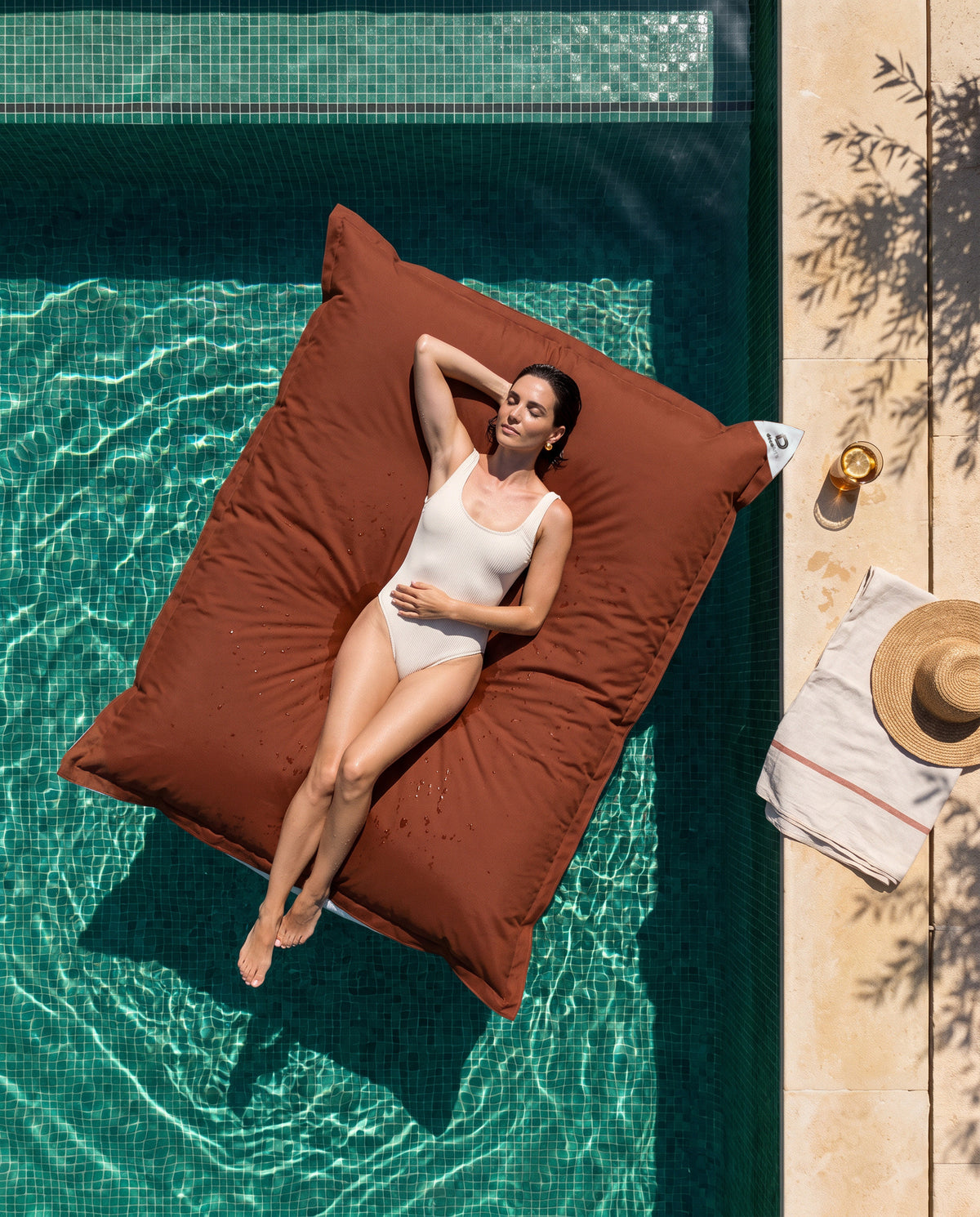 Pouf gigante galleggiante terracotta Deco Arts su una piscina a mosaico verde, vista dall'alto, donna sdraiata al sole